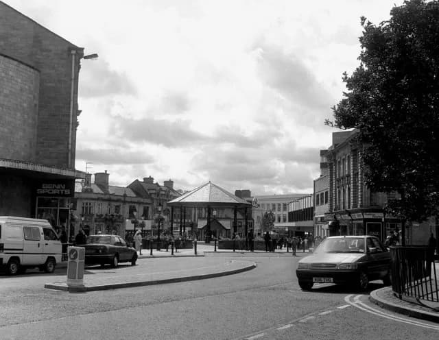 A black and white street scene with buildings flanking a road that curves around a central gazebo, under a cloudy sky.