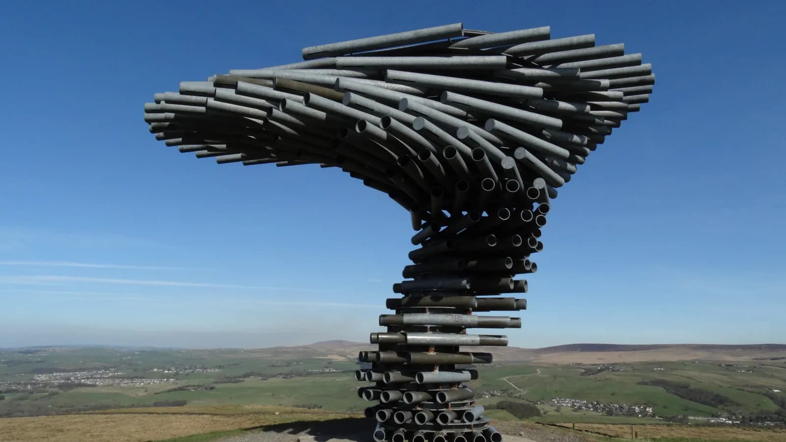 A large, metal, tree-shaped sculpture, composed of numerous pipes, stands on a hillside overlooking a green valley with scattered buildings under a clear blue sky.